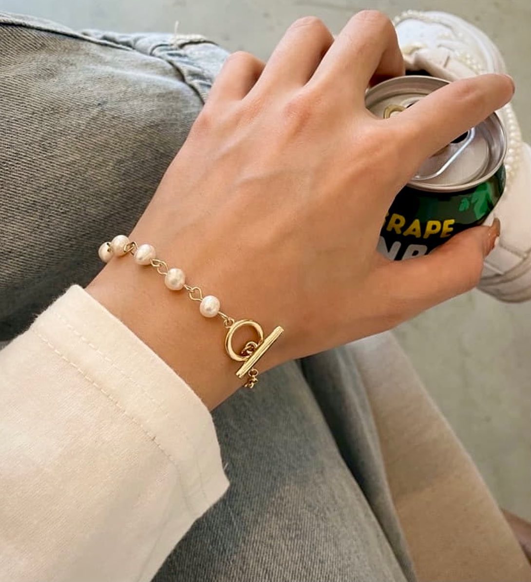 A gold-tone toggle clasp bracelet with white faux pearls worn on a person's wrist, shown while they hold a can of grape soda. The person is wearing a cream-colored long-sleeve top and gray jeans.