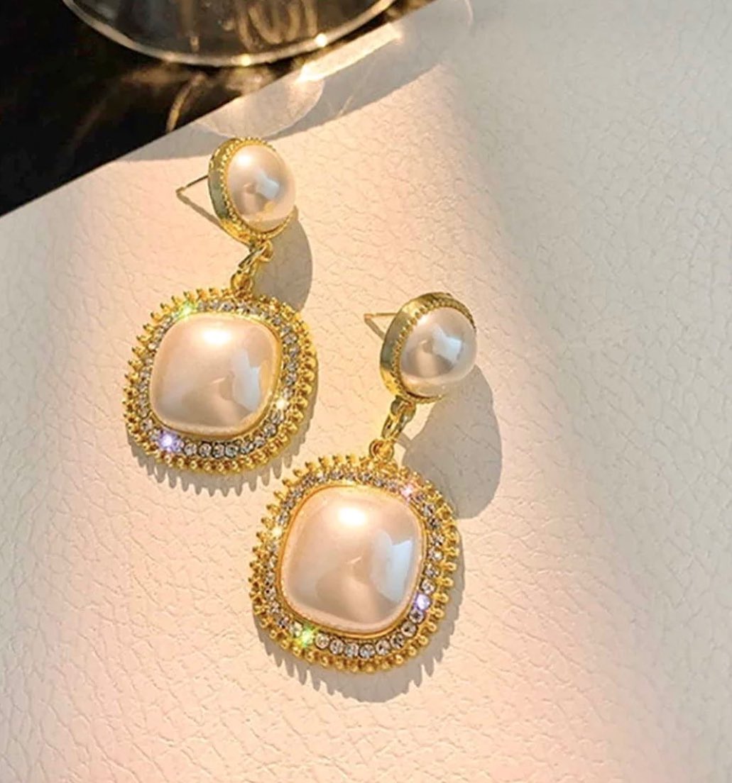 A pair of gold-tone drop earrings with large pearlescent oval center stones and smaller round pearl-like studs, displayed on a light textured surface.