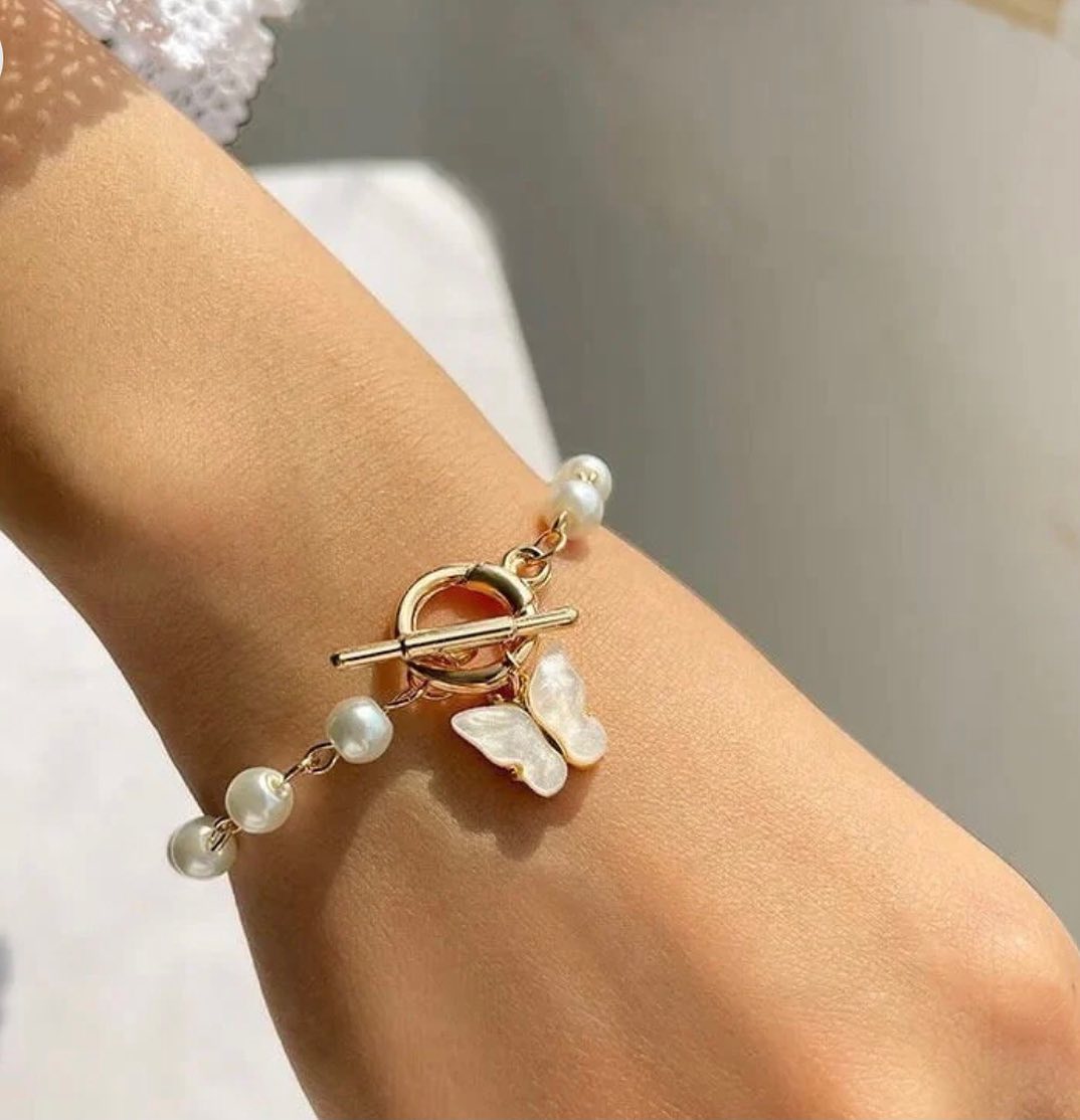 A close-up of a gold-tone toggle bracelet with white freshwater pearls and a butterfly charm, worn on a person's wrist.