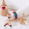 A set of five gold-toned statement rings displayed on a white textured fabric background, including a snake ring, a heart ring on a cone display, and colorful gemstone bands.