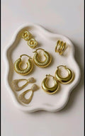 A set of gold-toned jewelry pieces arranged on a white scalloped tray against a light neutral background. The set includes hoop earrings, drop earrings, stud earrings, and a ring.