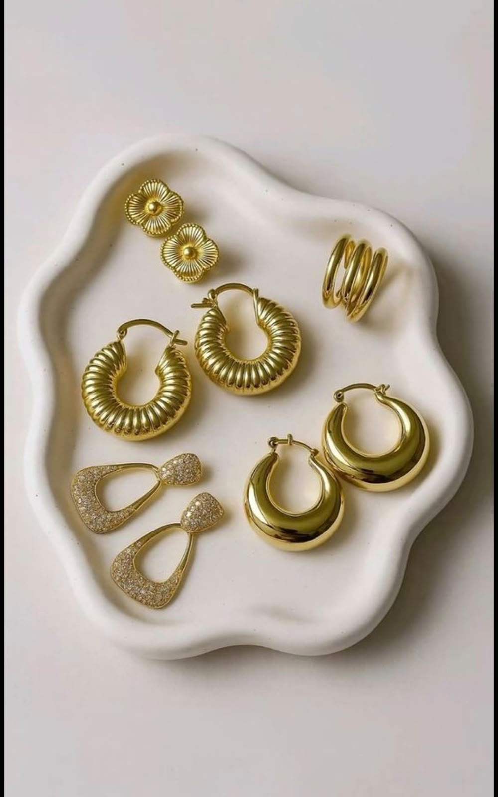 A set of gold-toned jewelry pieces arranged on a white scalloped tray against a light neutral background. The set includes hoop earrings, drop earrings, stud earrings, and a ring.