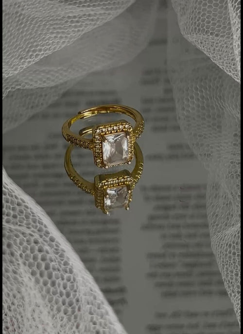 A gold ring with a square clear gemstone and small surrounding stones, displayed on a reflective surface with white sheer fabric draped around it.
