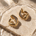 Gold abstract huggie hoop earrings with pavé-set clear stones, displayed on a textured beige stone slab with water ripples and dappled sunlight.