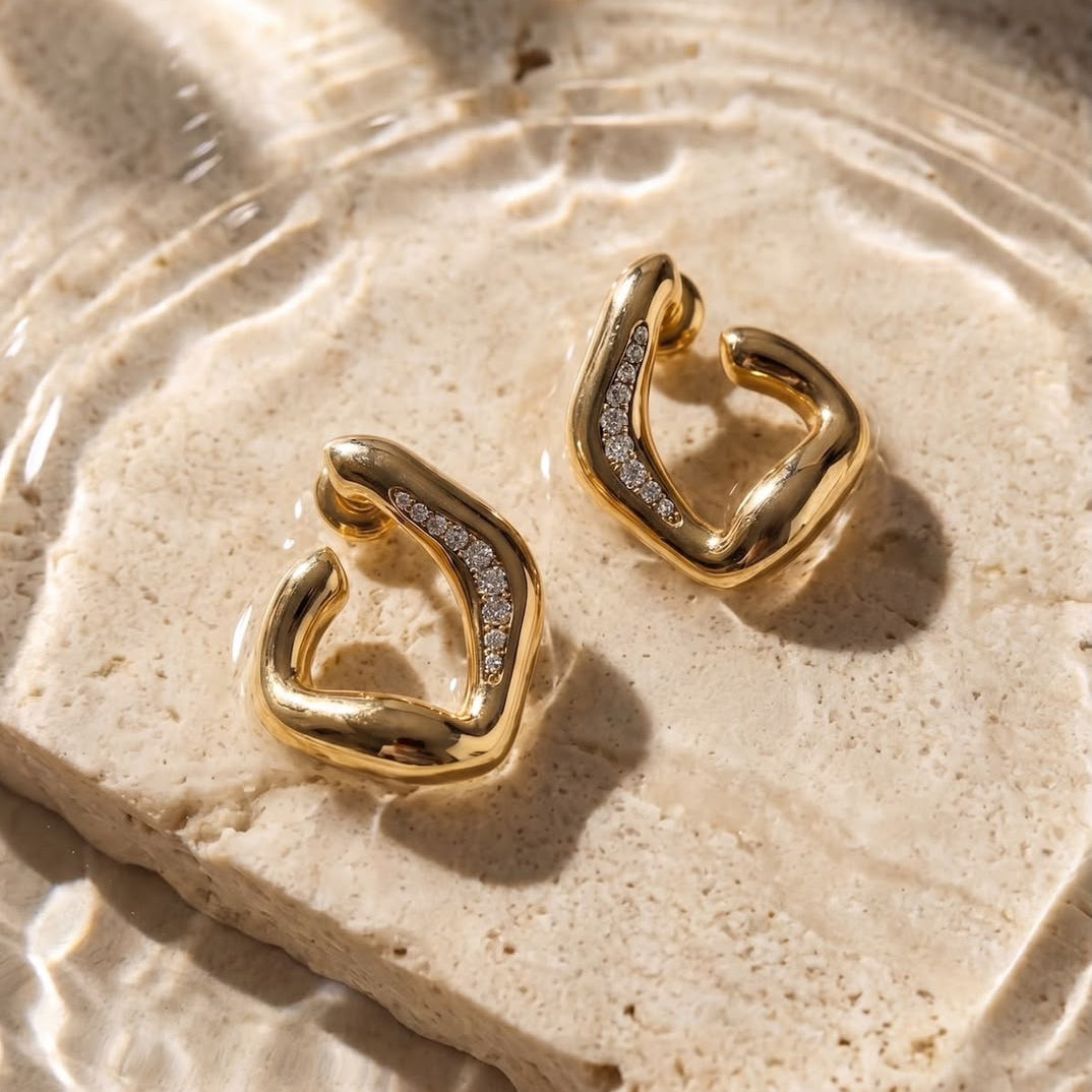 Gold abstract huggie hoop earrings with pavé-set clear stones, displayed on a textured beige stone slab with water ripples and dappled sunlight.